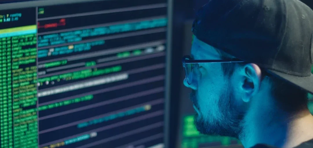 A person with glasses and a cap is focused on a computer screen displaying lines of code and data in green and blue hues. The screen represents DNS security and monitoring, highlighting the importance of preventing DNA attacks and protecting against website hijacking and cyber crime.